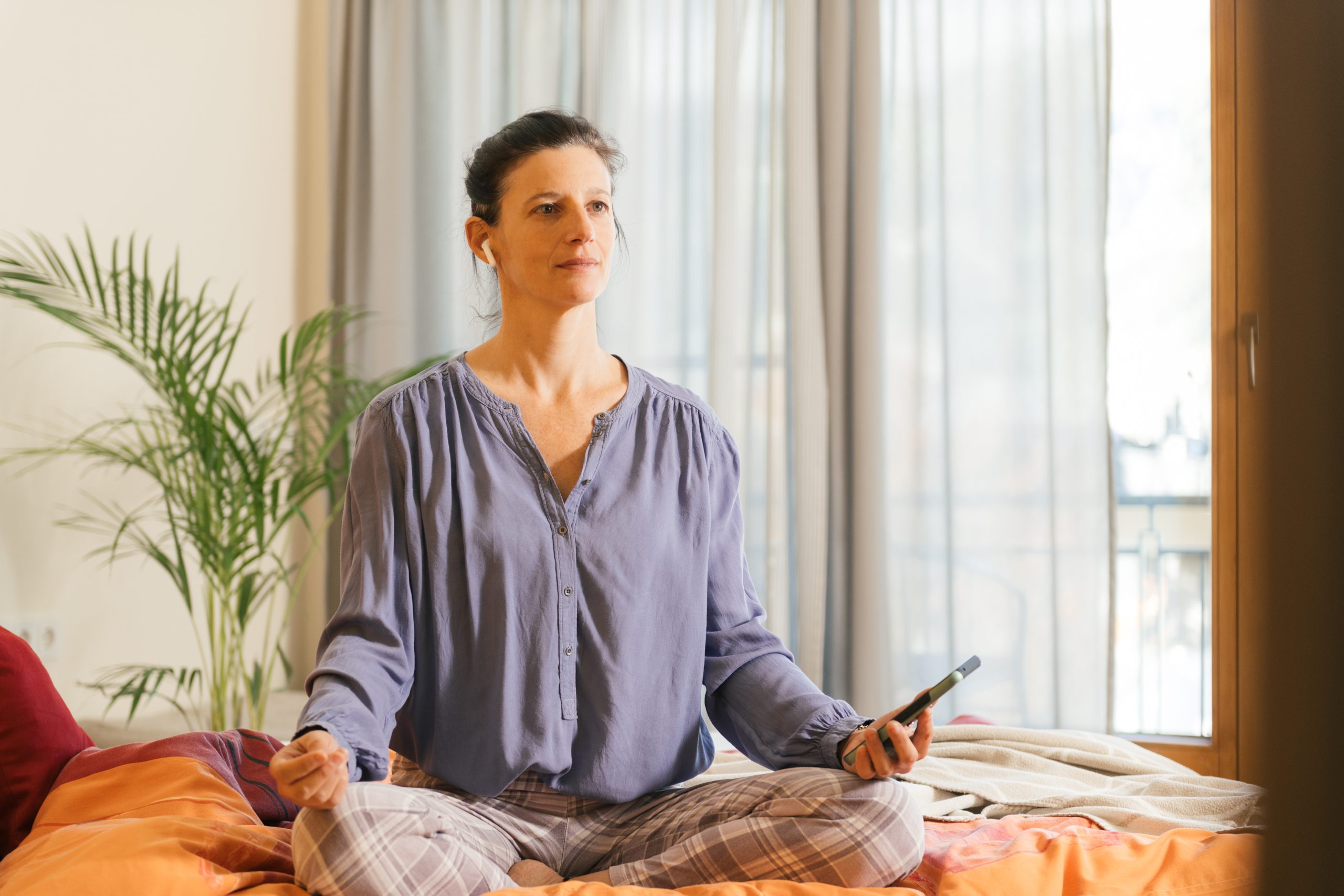 Eine Person im Meditationssitz praktiziert Energiearbeit und hält ein Smartphone, umgeben von Pflanzen in entspannter Atmosphäre.