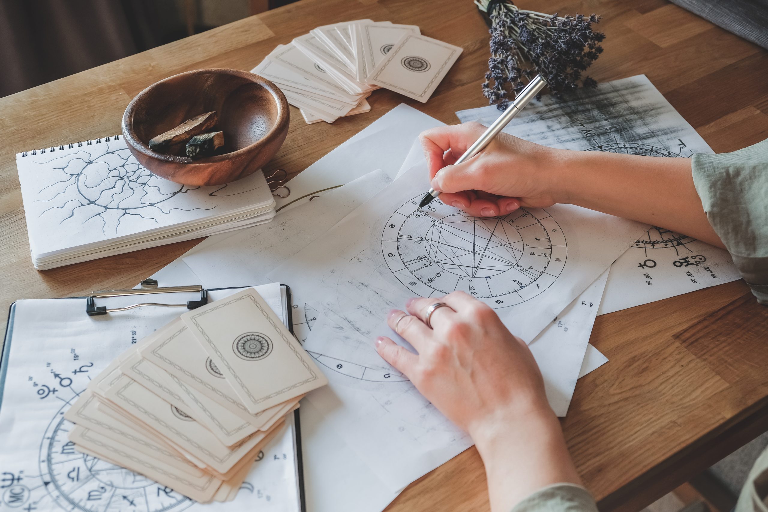 Person erstellt astrologische Diagramme f&uuml;r Energiearbeit mit Karten und Notizen auf einem Holzschreibtisch.